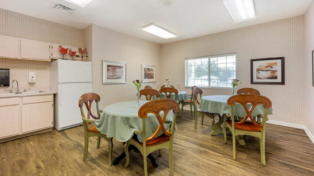 A small dining area with round tables covered in green cloths, wooden chairs, and simple decor. A window lets in natural light, and there are framed pictures and a refrigerator along one wall.