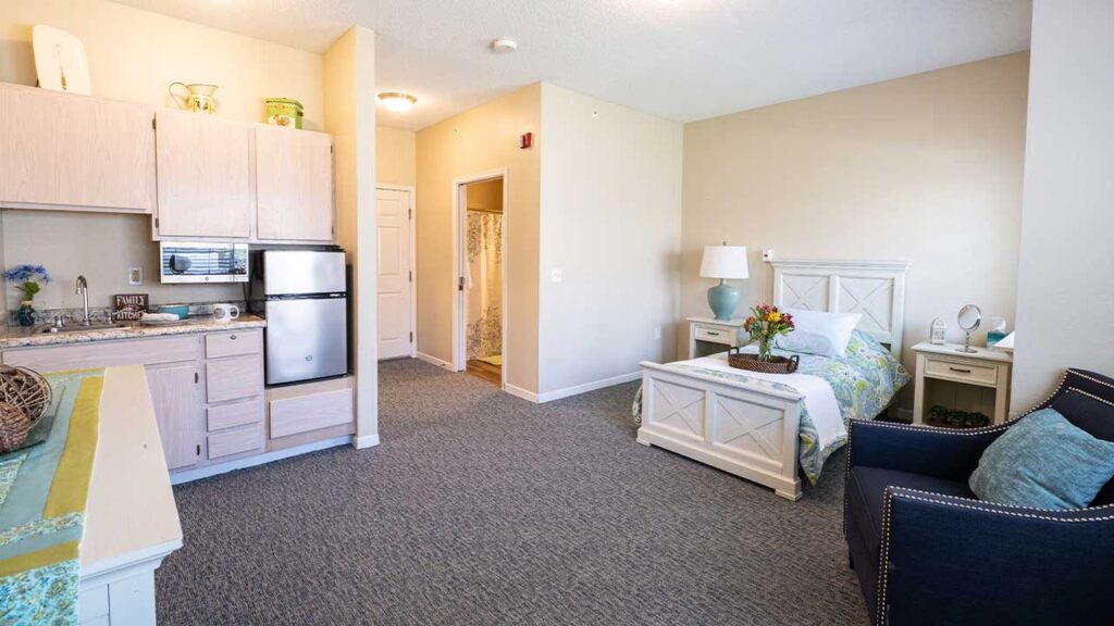 A cozy, well-lit studio apartment with beige walls and carpeted floor. It features a kitchenette with light wooden cabinets, a small stainless steel refrigerator, a bed with white bedding, a nightstand with a lamp, an armchair, and a small table with flowers. A door leads to a bathroom.