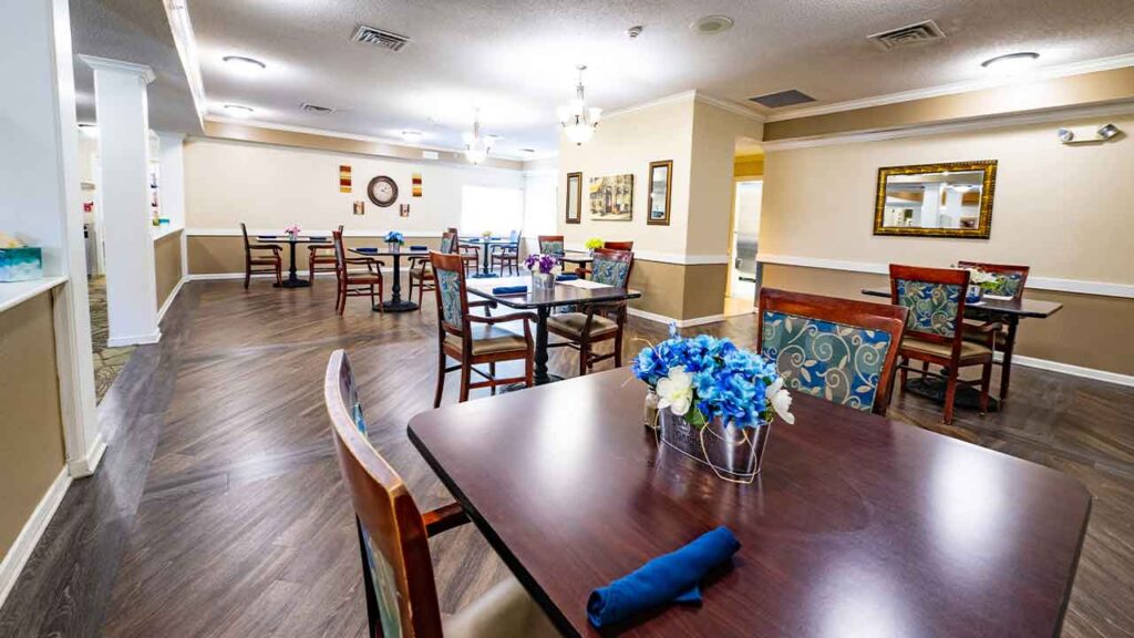 A cozy dining area featuring neatly arranged tables covered with dark wooden tops. Each table has napkins and a small floral centerpiece. The room has a warm ambiance with decorative wall art, mirrors, and soft lighting from chandeliers.
