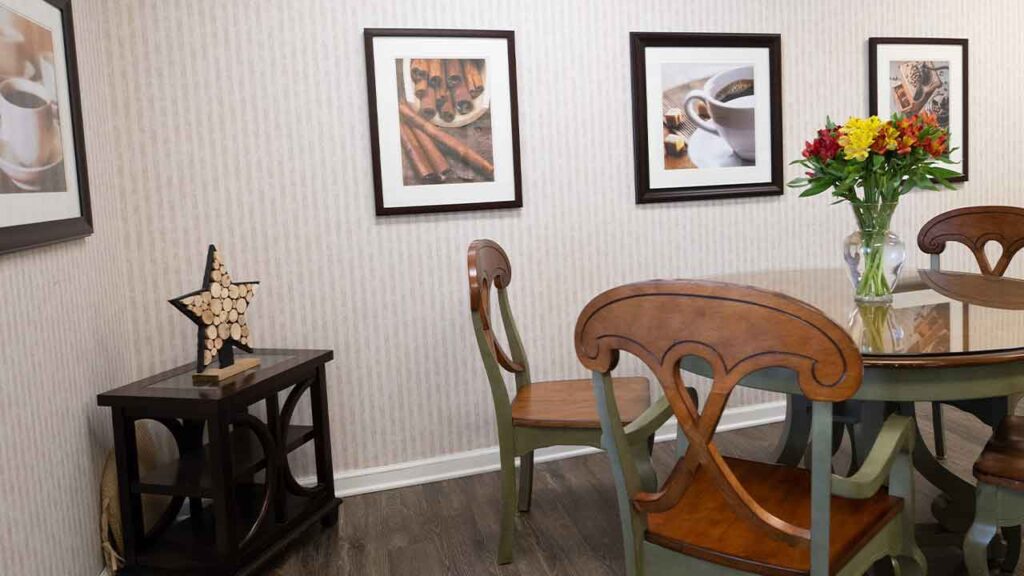 A dining room with a round wooden table, surrounded by four chairs. A vase with colorful flowers sits on the table. The walls are adorned with framed pictures of coffee-themed artwork. A small side table with a wooden star decoration is against the wall.