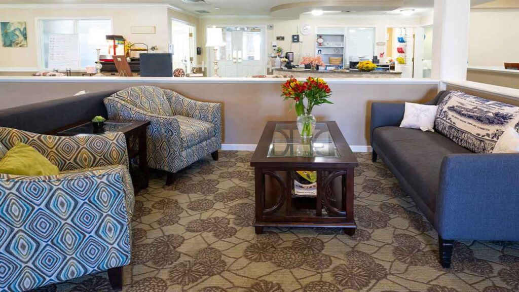 A cozy living room with patterned armchairs, a dark grey sofa, and a glass-top wooden coffee table holding a vase with red and yellow flowers. The room has a patterned carpet, neutral walls, and a counter with various items in the background.