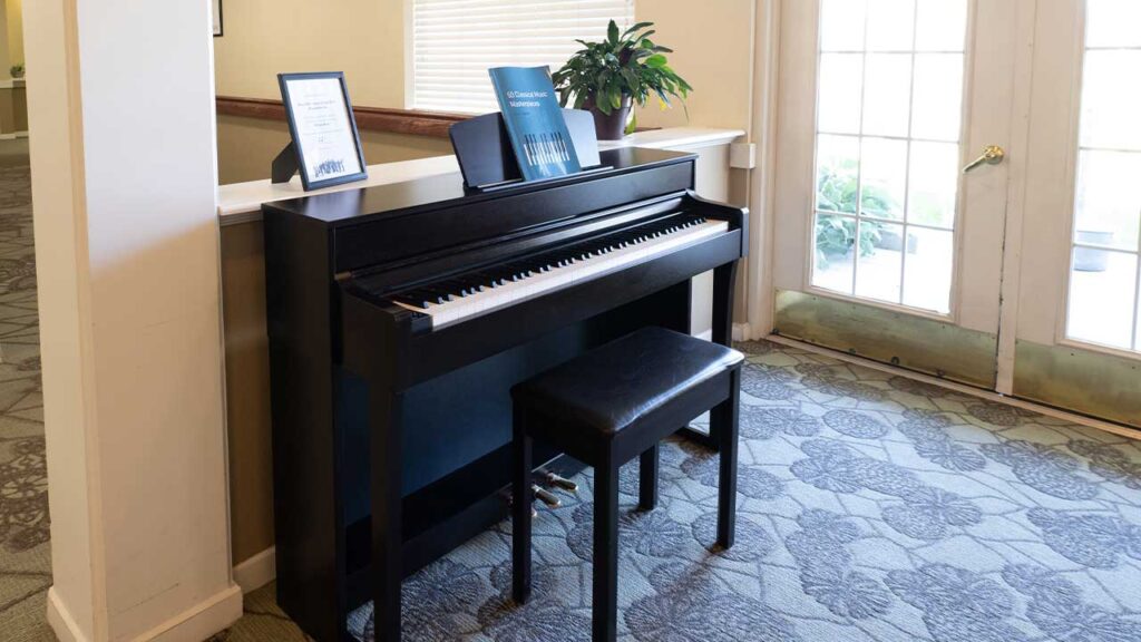 A black digital piano with a matching bench is placed against a wall near a set of glass French doors. Sheet music is open on the piano's stand, and the room features a patterned carpet and a potted plant in the background.