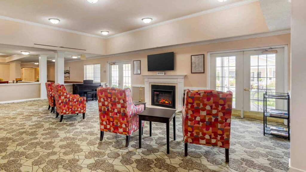 A cozy lounge area with patterned armchairs facing a fireplace and wall-mounted TV. Large windows and glass doors let in natural light, and the space has floral carpet and soft overhead lighting.