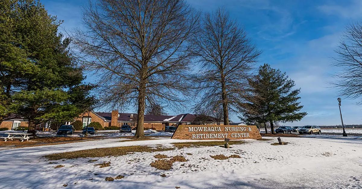 Sign for Moweaqua Nursing, Rehabilitation & Health Care Center amidst a snowy landscape with leafless trees and a clear blue sky. Nearby, several parked cars and a building are visible.