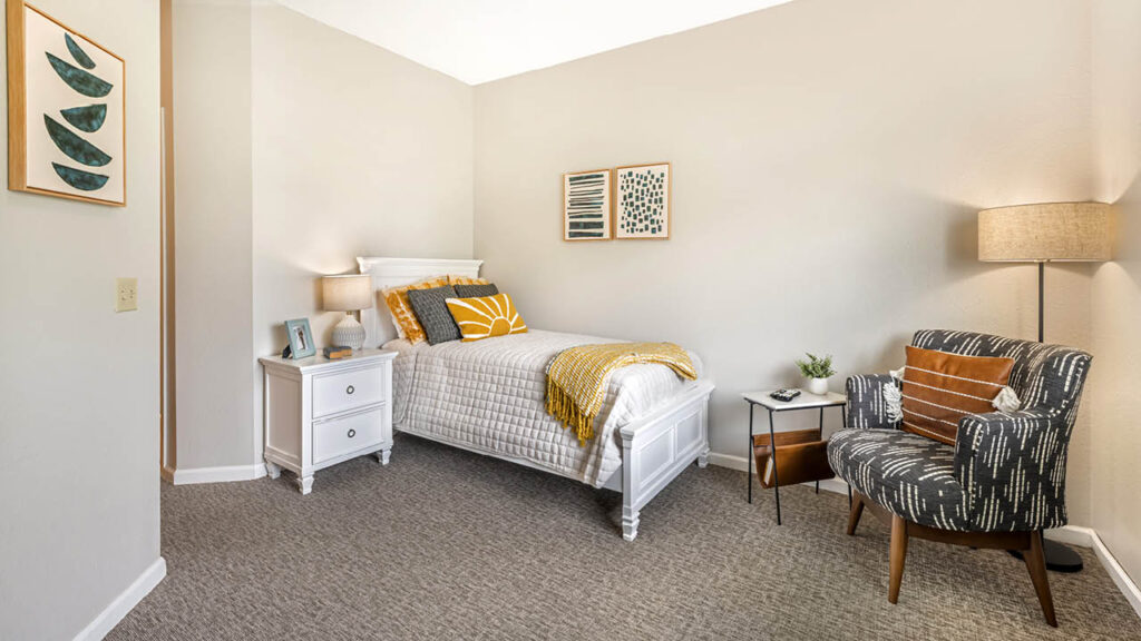A cozy bedroom with a single bed, white nightstand, patterned armchair, lamp, wall art, and light gray carpet. The decor features neutral tones with yellow and green accents.