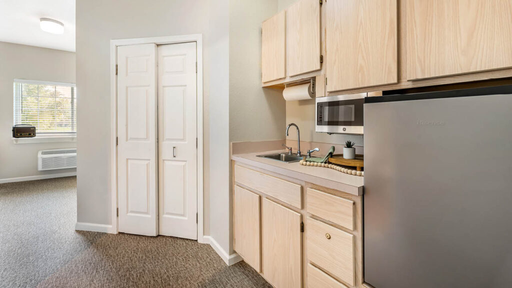 A small kitchenette with light wood cabinets, a sink, a microwave, a mini refrigerator, and a cutting board with a knife. An adjacent room with a window and an air conditioner is visible in the background.