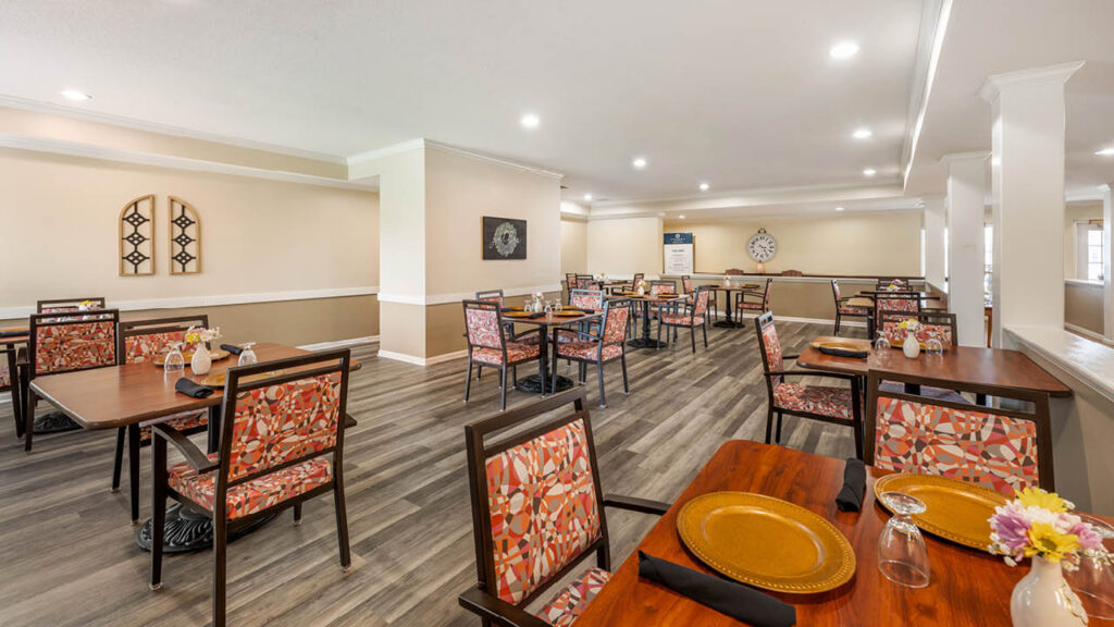 A spacious, brightly lit dining area with several tables set with plates, glasses, and floral centerpieces. The room features patterned chairs, wooden floors, and neutral-colored walls with decorative art pieces.