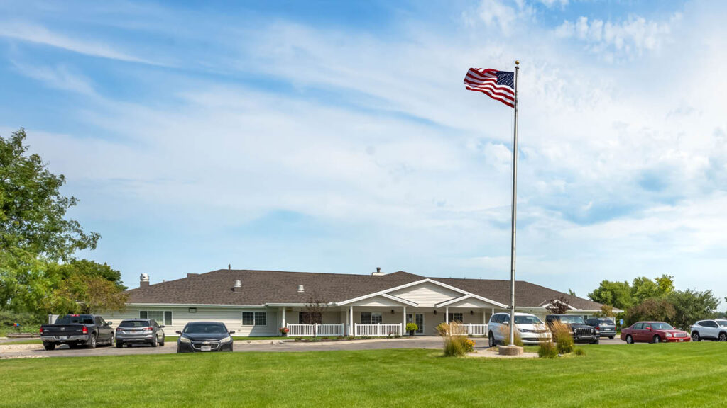 A single-story building with a covered entrance, several parked cars, and a large American flag flying on a flagpole, all set against a blue sky and grassy lawn.