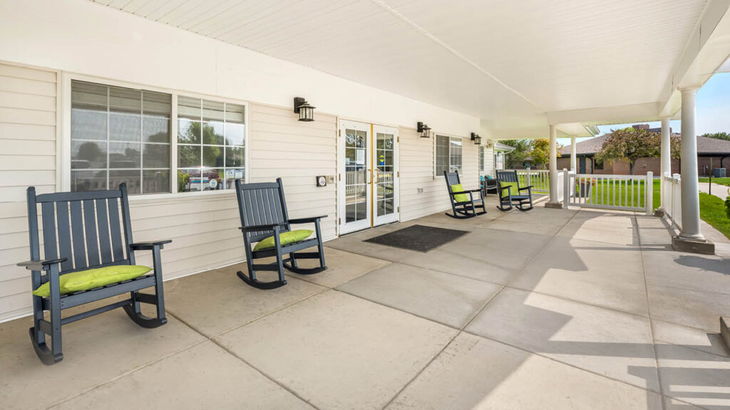 A wide covered porch with white railings and several dark wooden rocking chairs with green cushions, outside a light-colored building with double glass doors and windows. The porch overlooks a grassy area and parked cars.