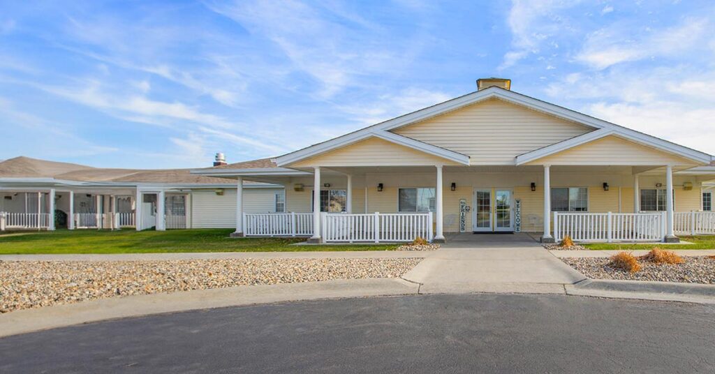 Single-story beige building with a wide covered front porch, white railings, and columns. A paved driveway leads to the entrance. There is a rock garden and grass lawn in front, under a blue sky with scattered clouds.