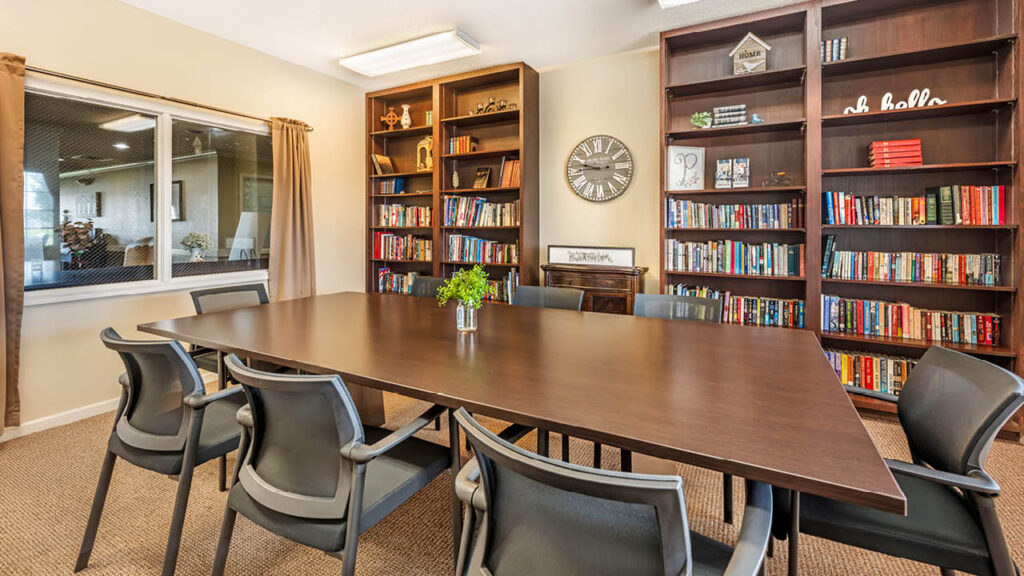 A conference room with a long wooden table, eight black chairs, bookshelves filled with books, a decorative wall clock, and a window with beige curtains. A small plant sits in the center of the table.