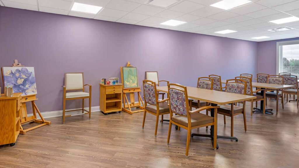 A bright room with purple walls, wooden floors, several tables and chairs, and art easels displaying paintings. Large windows let in natural light. The space appears to be set up for an art class or creative activity.