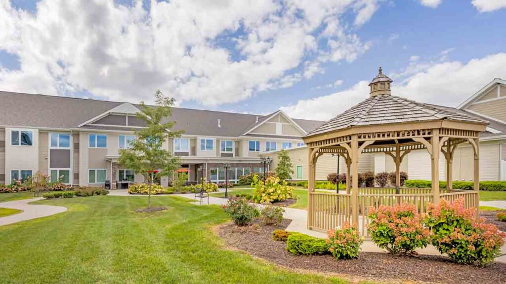 A landscaped courtyard with a wooden gazebo, green lawn, flower beds, and trees in front of a large, two-story building under a partly cloudy sky.