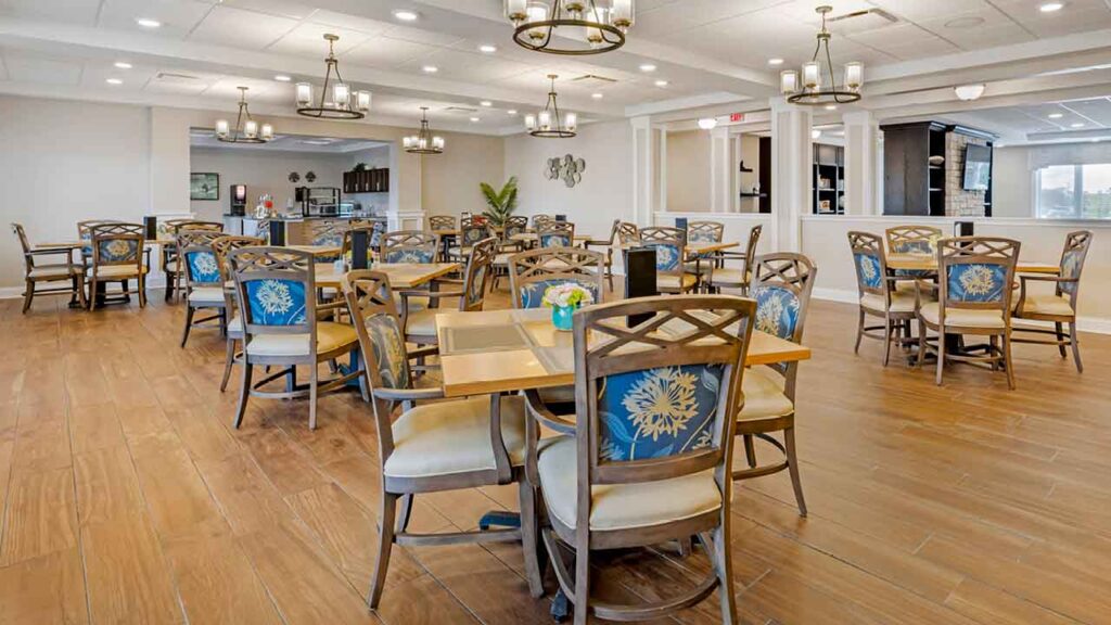 Spacious dining area with wooden floors, multiple square tables, and chairs with blue-patterned cushions. Modern light fixtures hang from the ceiling, and large windows let in natural light.