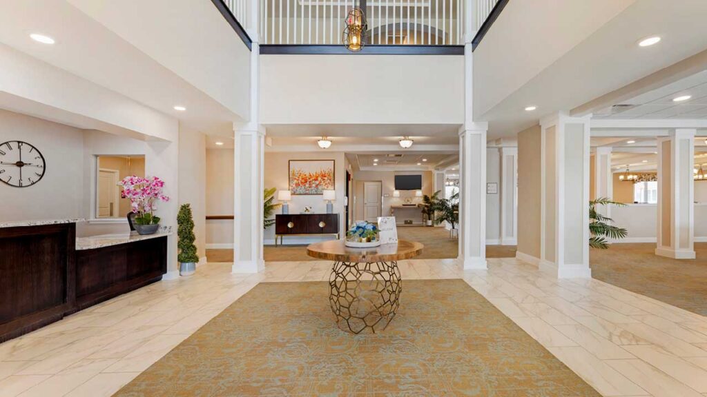 A spacious hotel lobby with a round wooden table in the center, beige and white decor, bright lighting, a clock on the left wall, flowers, and a staircase with a white railing above.
