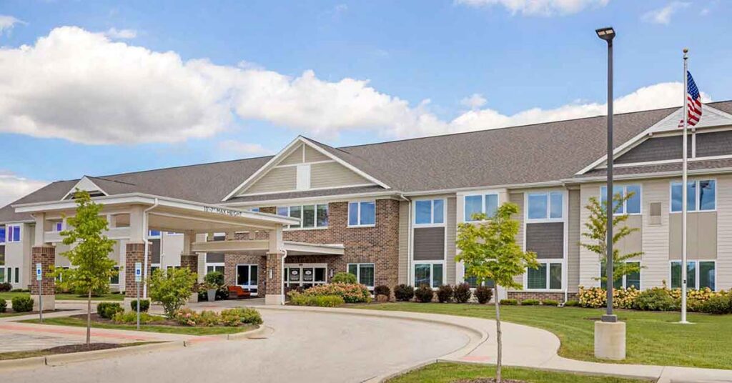 A modern, two-story building with a covered entrance, American flag, and landscaped greenery under a partly cloudy sky. The exterior features brick and siding, with large windows and a driveway in front.