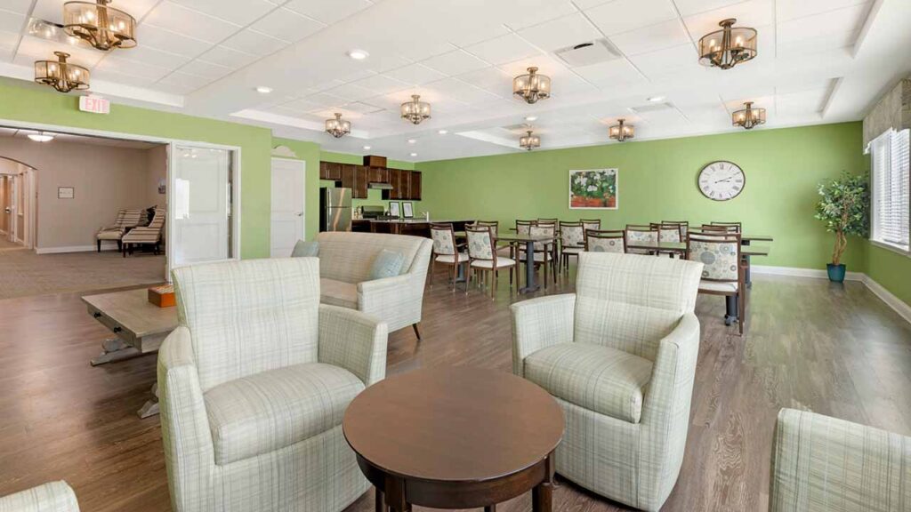 A bright, modern lounge with plaid armchairs, round tables, and dining tables on wood flooring. Green walls, a wall clock, large windows, and decorative lighting create a welcoming atmosphere.