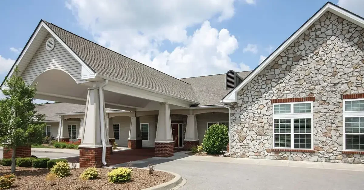 The image shows the Charlestown Senior Living Community, a single-story building with a wide driveway and a sheltered entrance. The facade features a mix of stone and brickwork, with large windows and a gabled roof. Landscaped bushes and a tree are visible around the entrance. The sky is partly cloudy.