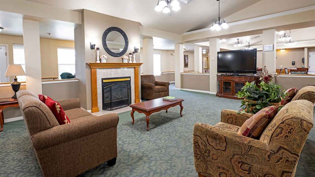 A cozy living room with beige walls and green carpet features a fireplace with a decorative mirror above it. Two brown armchairs and a sofa surround a wooden coffee table. A large TV stands in the background, and the room is well-lit with ceiling lights.