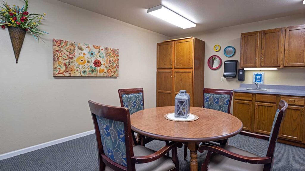 A cozy room with a round wooden table surrounded by four upholstered chairs. A decorative lantern sits on the table. The wall features a floral painting and a basket with colorful flowers. Wooden cabinets and a countertop are in the background.