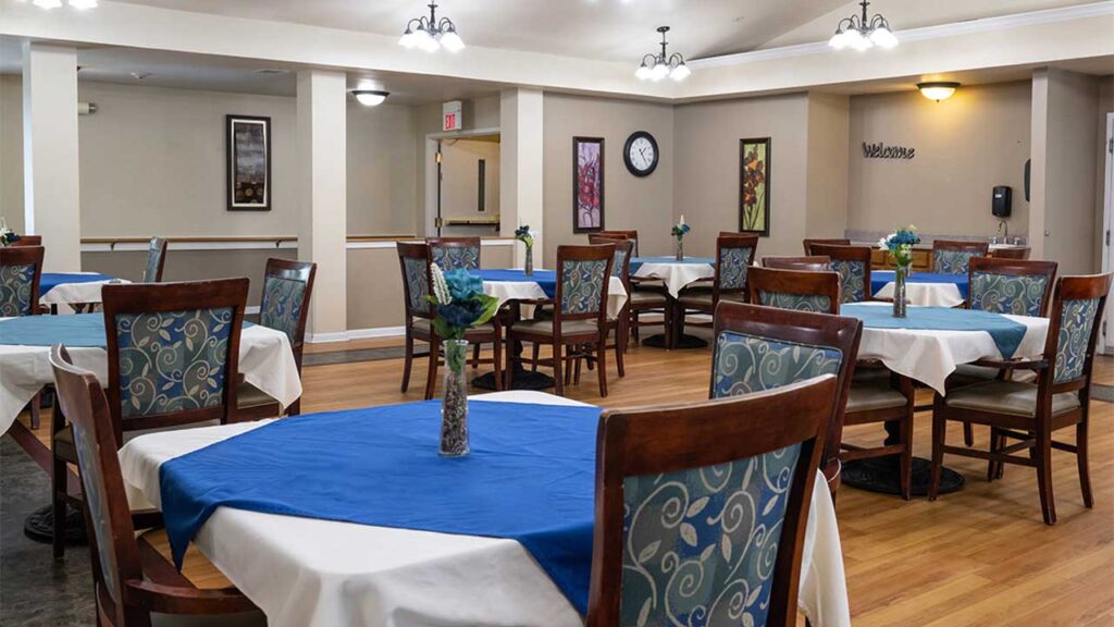 A dining area with round tables covered in white and blue cloths, each adorned with a small floral centerpiece. Wooden chairs with patterned cushions surround the tables. The walls are decorated with framed artwork, a clock, and a