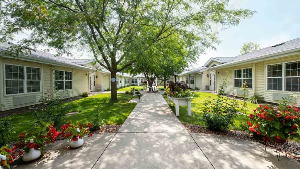 A serene courtyard with a concrete walkway flanked by lush green grass and manicured garden beds featuring vibrant red and white flowers. The area is shaded by large green trees and surrounded by single-story residential buildings with large windows.