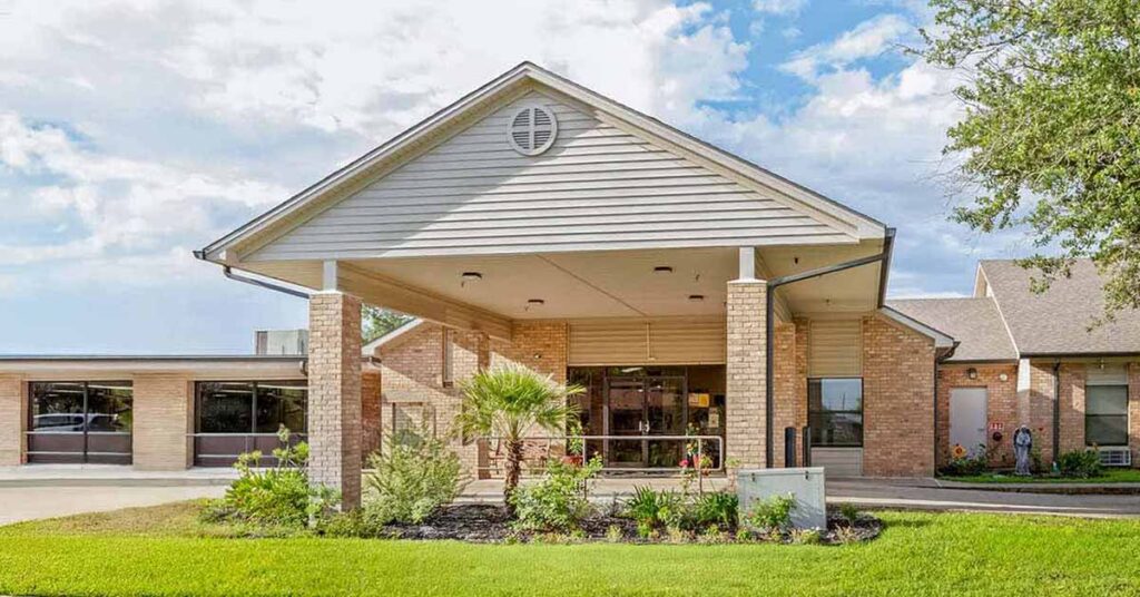 A single-story brick Nursing & Rehabilitation building with a covered entrance, landscaped garden, and large windows sits against a partly cloudy sky. A driveway runs in front of the entrance.