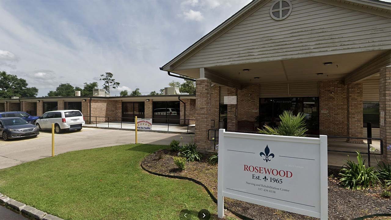 A single-story building with a brick exterior. A sign in front reads “Rosewood Nursing & Rehabilitation, Est. 1965.” There is a parking lot with a few cars and a covered entrance with plants on either side. The sky is partly cloudy.