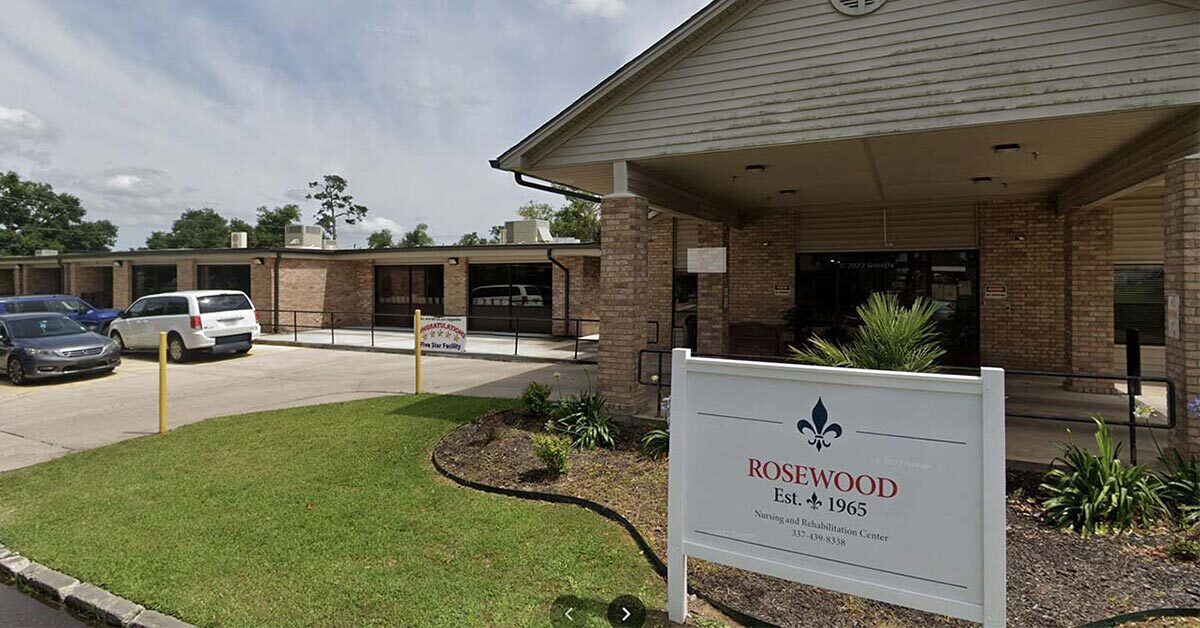 The entrance of the brick Rosewood building, established in 1965, features a sign with a fleur-de-lis symbol, welcoming visitors to this nursing and rehabilitation center. The landscaped area leads to a parking lot filled with cars beneath the partly cloudy sky.