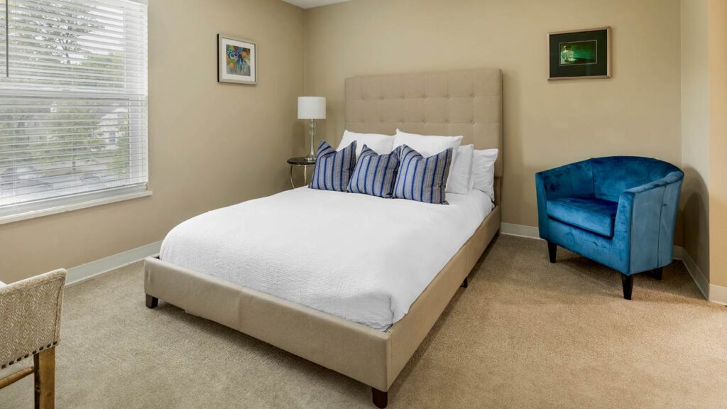 A clean and modern bedroom in a senior living space, featuring beige walls and carpet. It includes a beige upholstered bed with white bedding and multiple blue-striped accent pillows. A blue armchair sits in the corner, accompanied by framed artworks on the walls. A large window lets in natural light.