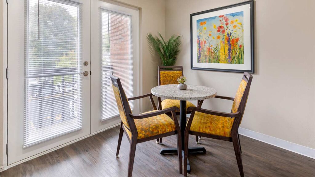 A cozy senior living dining area with a round table featuring a small plant centerpiece and surrounded by four yellow patterned chairs. French doors with blinds lead outside, and a colorful floral painting adorns the beige wall. A green potted plant sits in the corner.