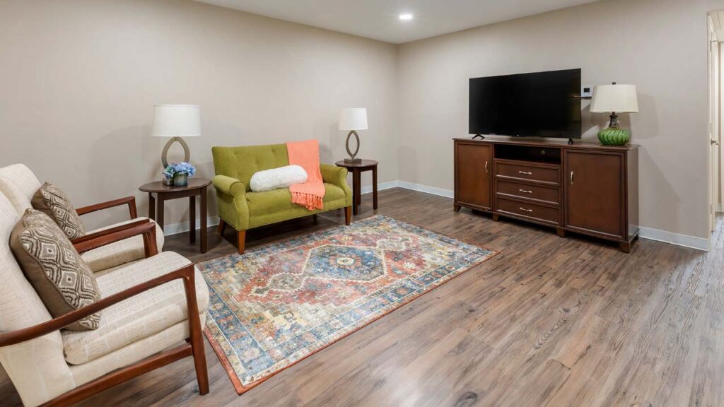 A cozy senior living room features a green loveseat with an orange throw and white cushion, two beige armchairs with patterned pillows, a colorful area rug, two table lamps, a wooden TV stand with a flat-screen TV, and wood flooring.