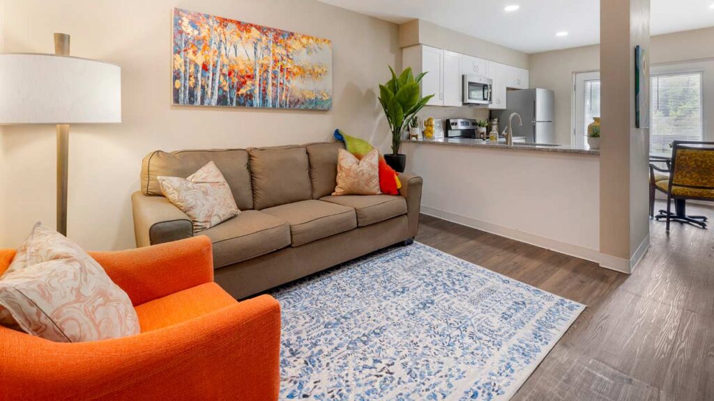 A modern senior living room with a gray sofa, multicolored cushions, an orange armchair, and a blue patterned rug. A lamp and vibrant artwork featuring trees adorn the beige wall. To the right, a kitchen area with stainless steel appliances and a potted plant is visible.