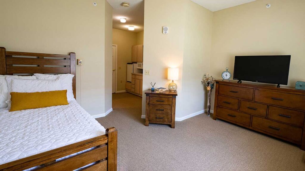 A cozy bedroom features a wooden bed with white bedding and a yellow accent pillow. Next to the bed is a wooden nightstand with a lamp and small items. Across the room is a wooden dresser with a TV on top, and decorative items on the side. A doorway leads to another room.