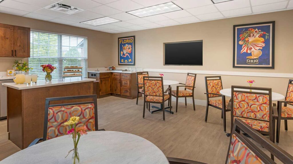 A bright, modern dining area with several round tables and patterned chairs, a kitchenette with drinks and snacks, large windows, wall art, and a flatscreen TV on a beige wall.