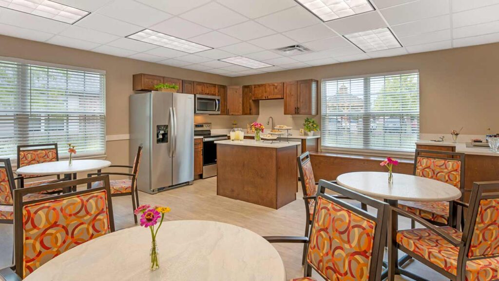 A modern communal kitchen with stainless steel appliances, wooden cabinets, and several round tables and chairs with orange patterned upholstery. Small vases with flowers are on the tables. Large windows let in natural light.