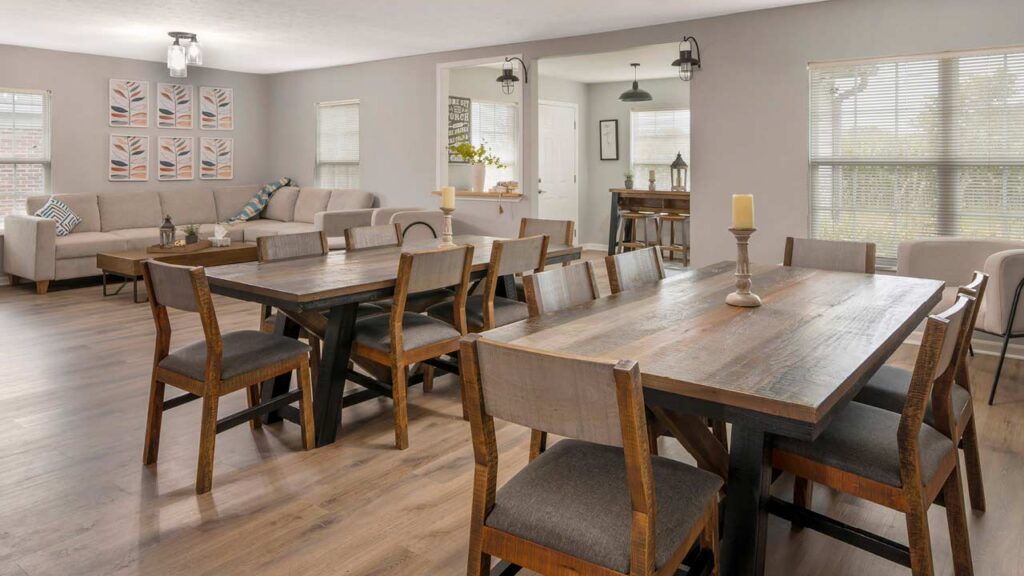 Open-plan living and dining area with two wooden tables, ten chairs, a beige sectional sofa, candles, decorative wall art, and large windows letting in natural light.