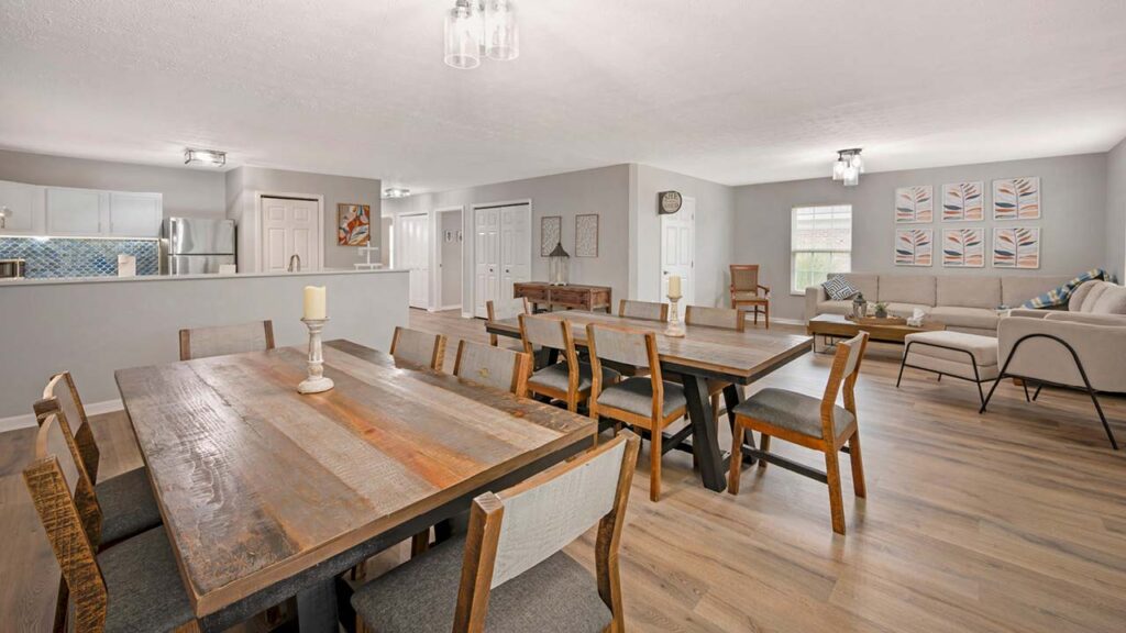 Open-concept dining and living room with wooden tables and chairs, neutral-toned couches, light wood floors, and modern light fixtures. The kitchen is visible in the background with white cabinets and stainless steel appliances.