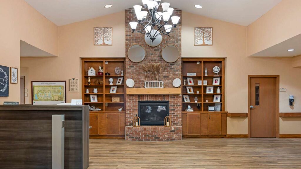 A cozy lobby with a brick fireplace, wooden shelves filled with framed photos and decor, a central chandelier, and warm-toned walls. A reception desk is on the left and a closed wooden door on the right.