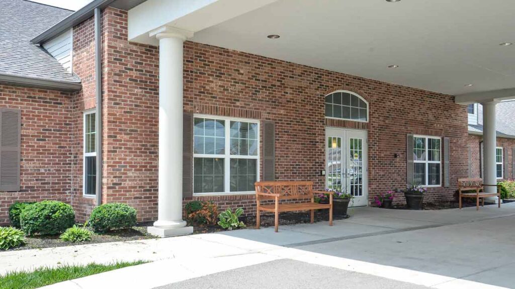 A brick building with white-framed windows, a white front door, benches, potted plants, and neatly trimmed bushes under a covered entrance.