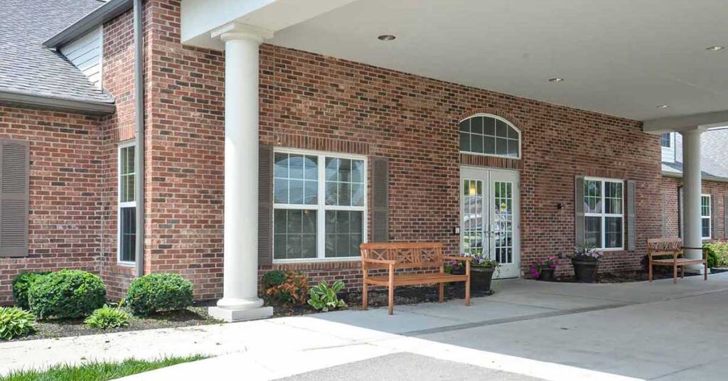 A red brick building with white trim, tall windows, two wooden benches, potted plants, and shrubs along the sidewalk under a covered entrance.