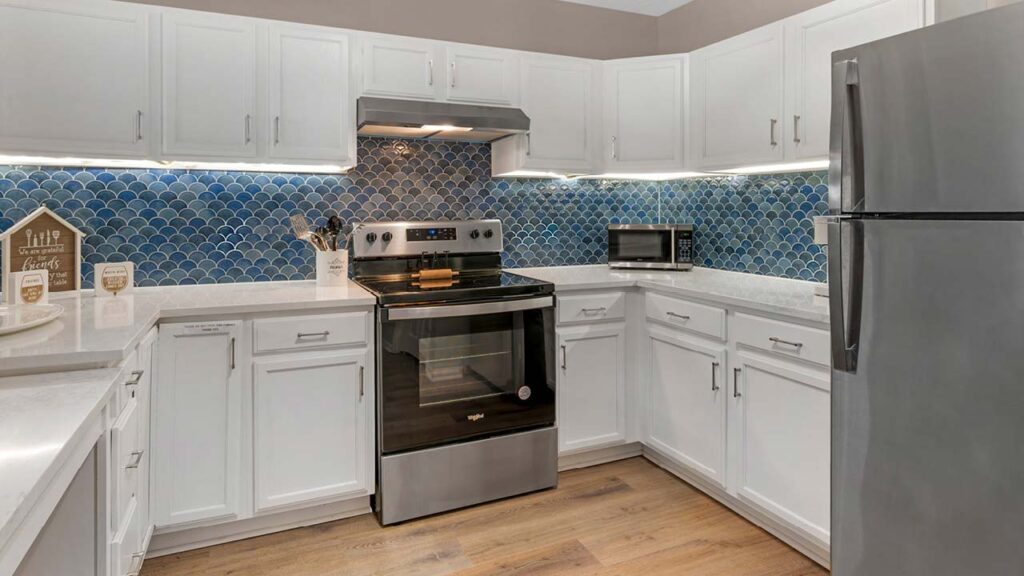 Modern kitchen with white cabinets, stainless steel appliances, blue fish scale tile backsplash, and light wood flooring. A stove, microwave, utensils, and decorative items are visible on the countertops.