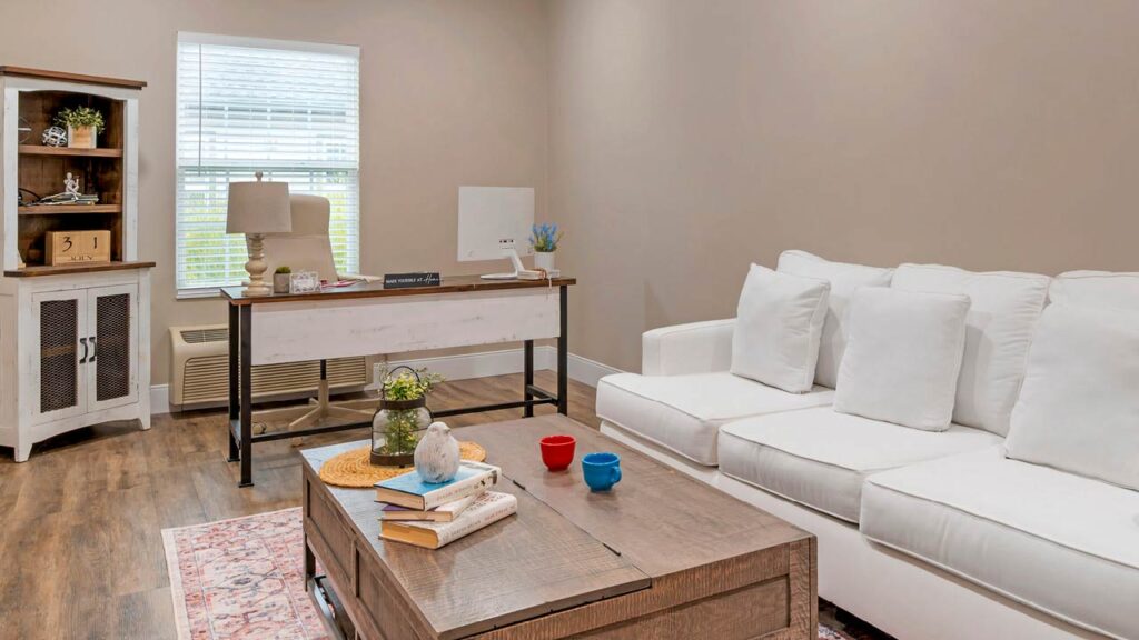A bright, cozy home office with a white desk, lamp, and computer by a window; a white sofa, a wooden coffee table with books and cups, and a bookshelf in the corner on wood flooring.