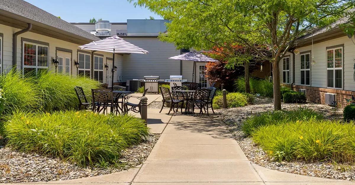 A peaceful outdoor courtyard featuring metal patio tables and chairs under striped umbrellas. The area is surrounded by well-maintained grass, small shrubs, and trees, set between two single-story buildings.