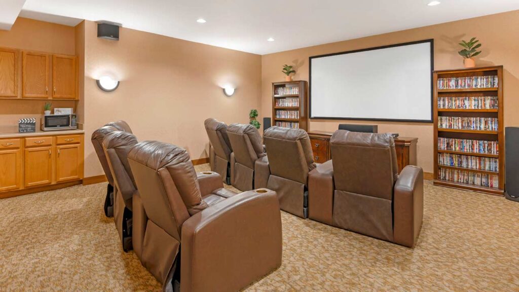 A cozy home theater room with six brown recliner chairs facing a large projector screen. Shelves filled with DVDs line the back wall, and built-in wooden cabinets are on the left. Warm lighting creates a comfortable atmosphere.