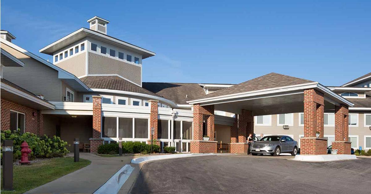 A modern, multi-story building with red brick and beige siding features a covered entrance with a driveway. Two people stand near a parked car under the entrance. The sky is clear and blue.