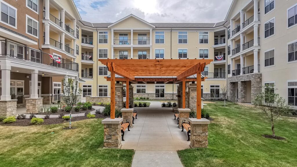 A spacious courtyard of a modern senior living apartment complex with a wooden pergola in the center, surrounded by benches and landscaped greenery. The four-story buildings have balconies, and an American flag is displayed on one of them. The sky is partly cloudy.