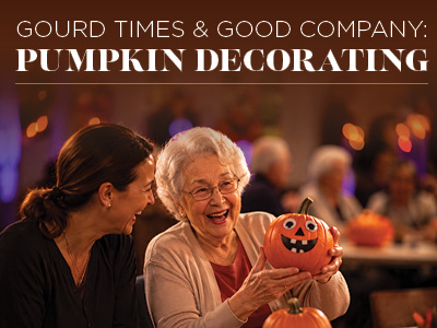 Two women smile and laugh while holding a decorated pumpkin. Warm lighting and blurred pumpkins in the background create a festive, cozy atmosphere. Text above reads, “Gourd Times & Good Company: Pumpkin Decorating.”.