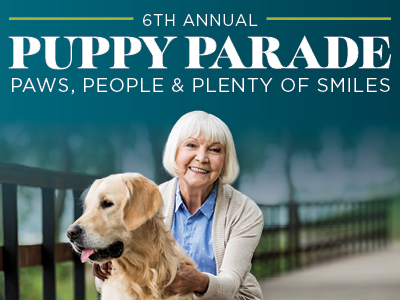 Smiling older woman with white hair sits outdoors on a wooden path, petting a golden retriever. Text above reads: “6th Annual Puppy Parade. Paws, People & Plenty of Smiles.”.