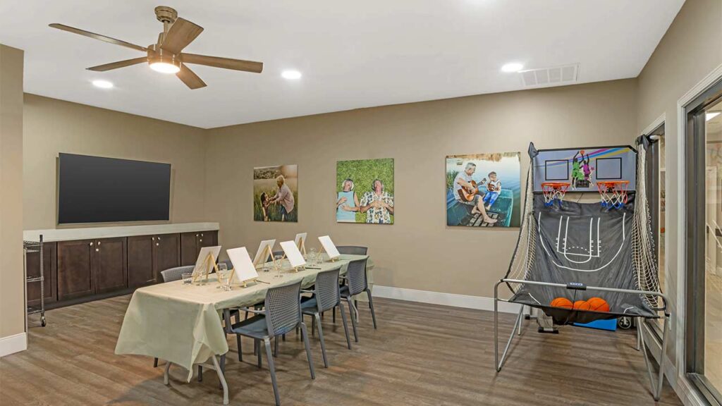 A bright assisted living room with wood floors features a long art table, wall-mounted TV, cabinets, family photos, and an indoor basketball arcade game with basketballs. A ceiling fan and vibrant lighting complete the welcoming space.